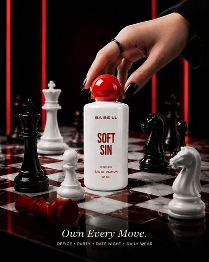 A hand with black nail polish reaches toward a white bottle of BA BE LL Soft Sin Eau de Parfum with a red cap, styled on a chessboard with chess pieces against a dark background with red vertical lights.