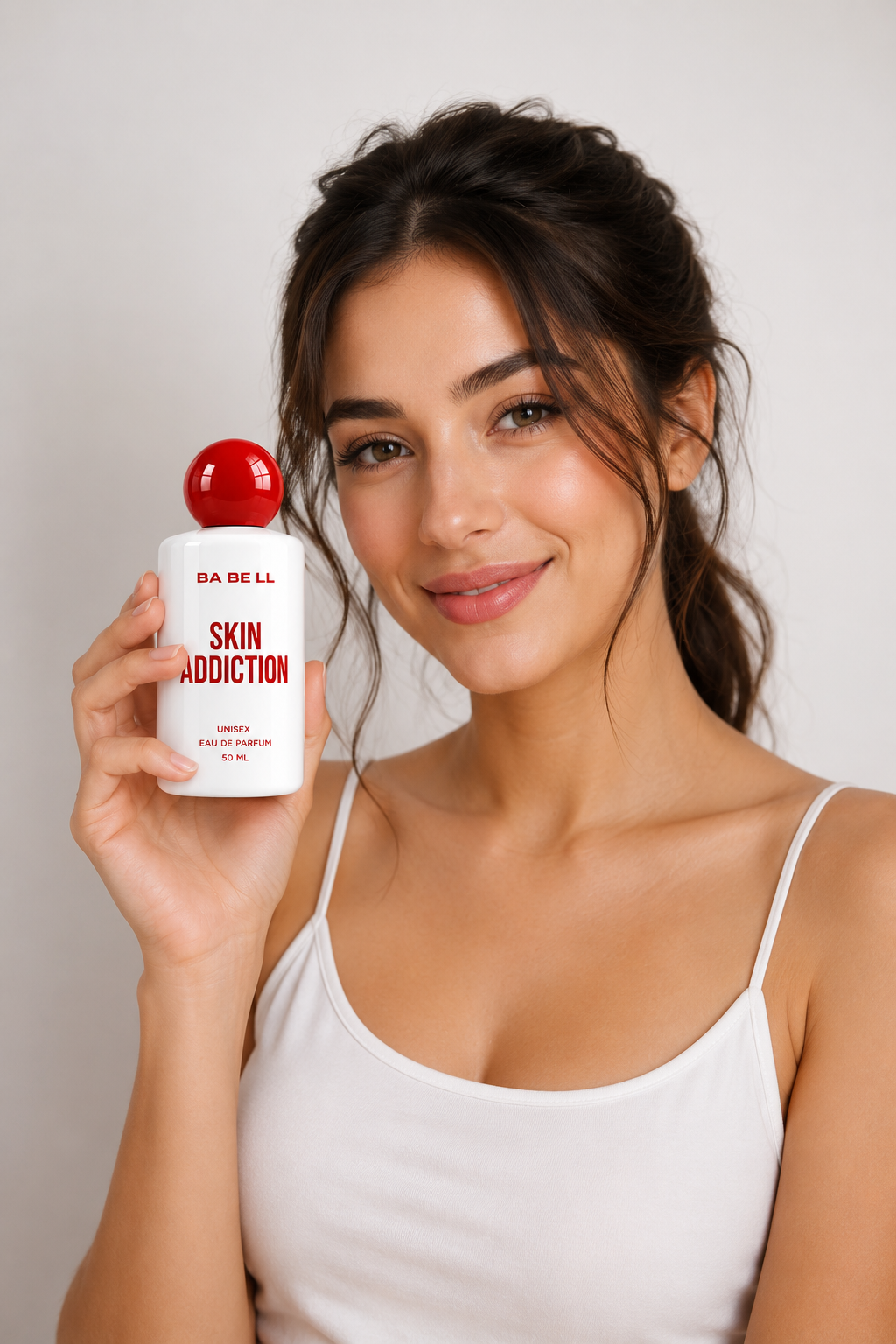A woman with dark hair tied back, wearing a white camisole, holds up a white bottle of BA BE LL Skin Addiction perfume with a red cap against a light gray background.