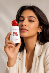 A woman in a cream blazer holds a white bottle of BA BE LL Loud Silence Eau de Parfum next to her face. The perfume bottle is white with bold red text and a red spherical cap, set against a warm beige background with soft shadows.