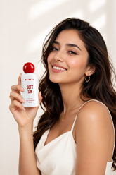 A woman holding a white bottle of BA BE LL Soft Sin perfume with a red cap, set against a warm, softly lit beige background with leaf shadows.